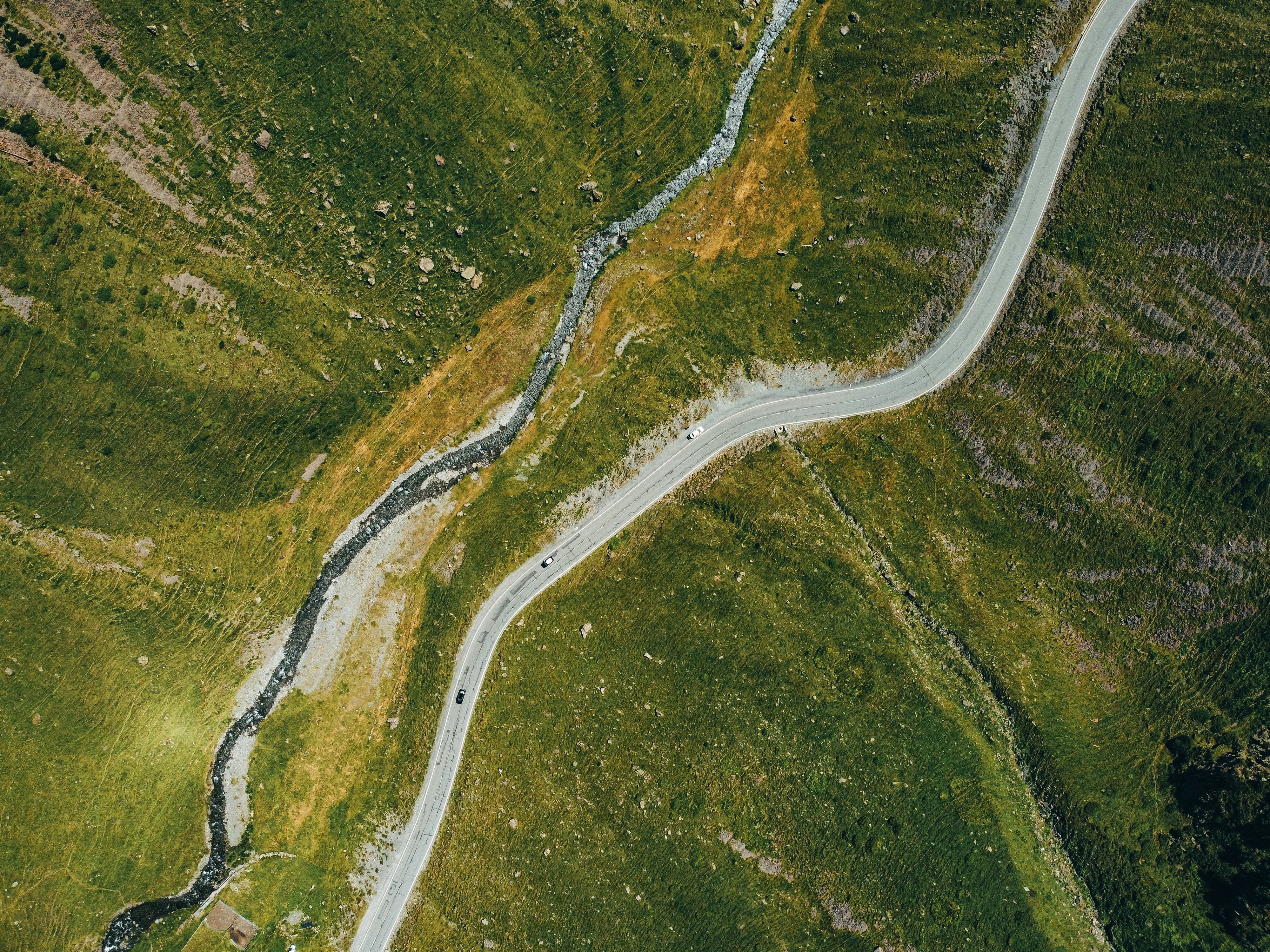 A rural mountain road running parallel to a stream cutting through a grassy landscape
